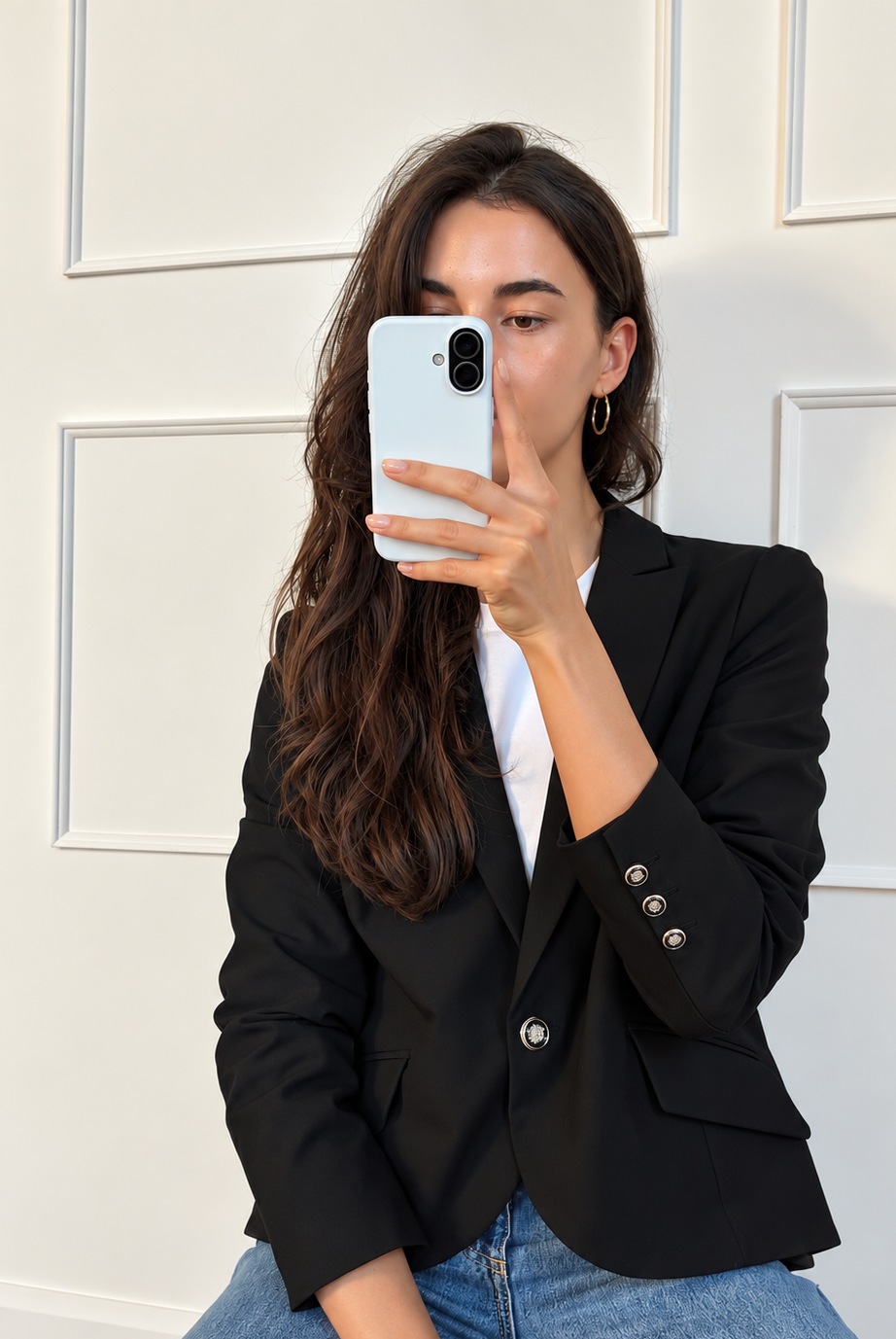 A woman taking a mirror selfie wearing a black blazer over a white t-shirt and blue jeans, holding a white smartphone.