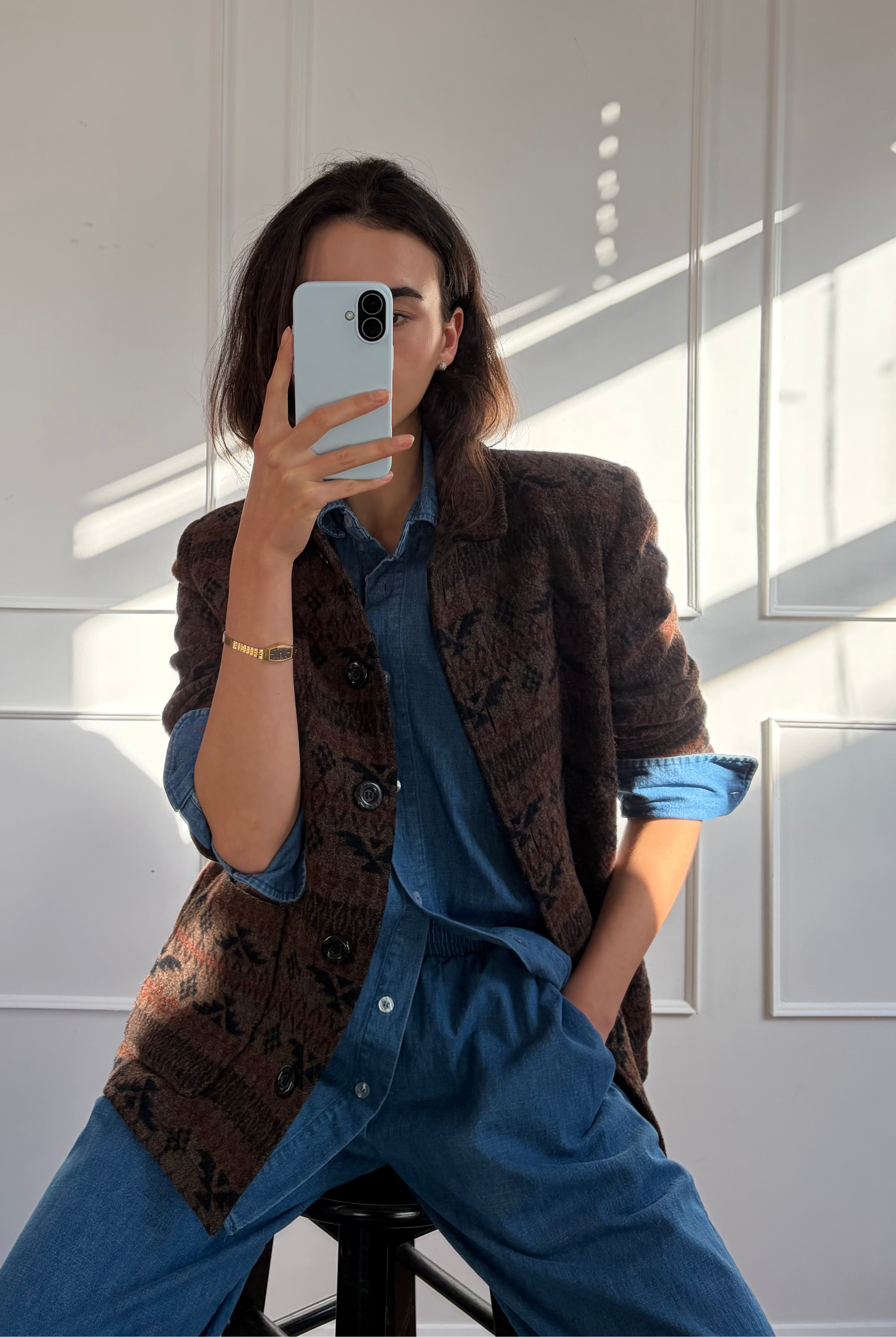 A person taking a mirror selfie in a room with white paneled walls and strong sunlight. They are wearing a brown patterned blazer over a blue button-up shirt and blue wide-leg pants, sitting on a black stool.