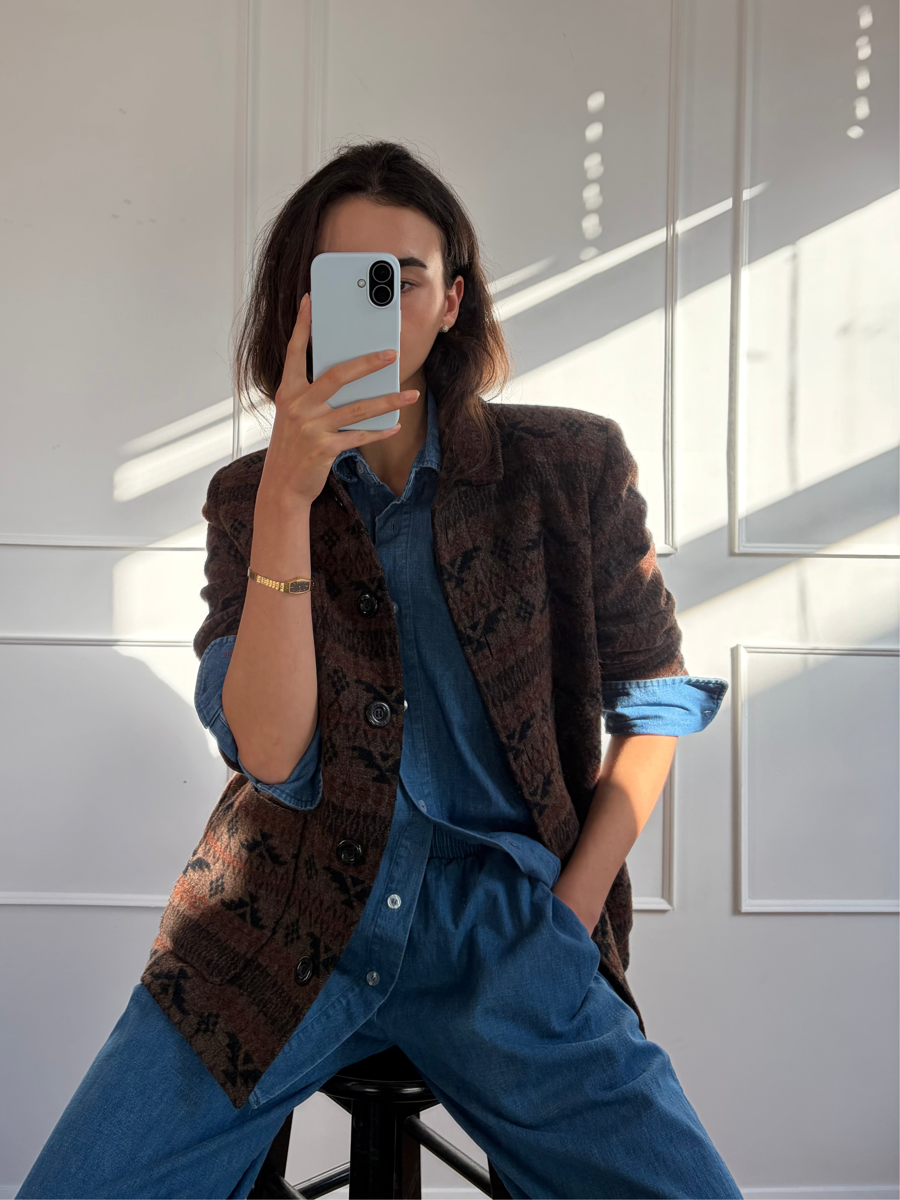 A person taking a mirror selfie in a room with white paneled walls and strong sunlight. They are wearing a brown patterned blazer over a blue button-up shirt and blue wide-leg pants, sitting on a black stool.