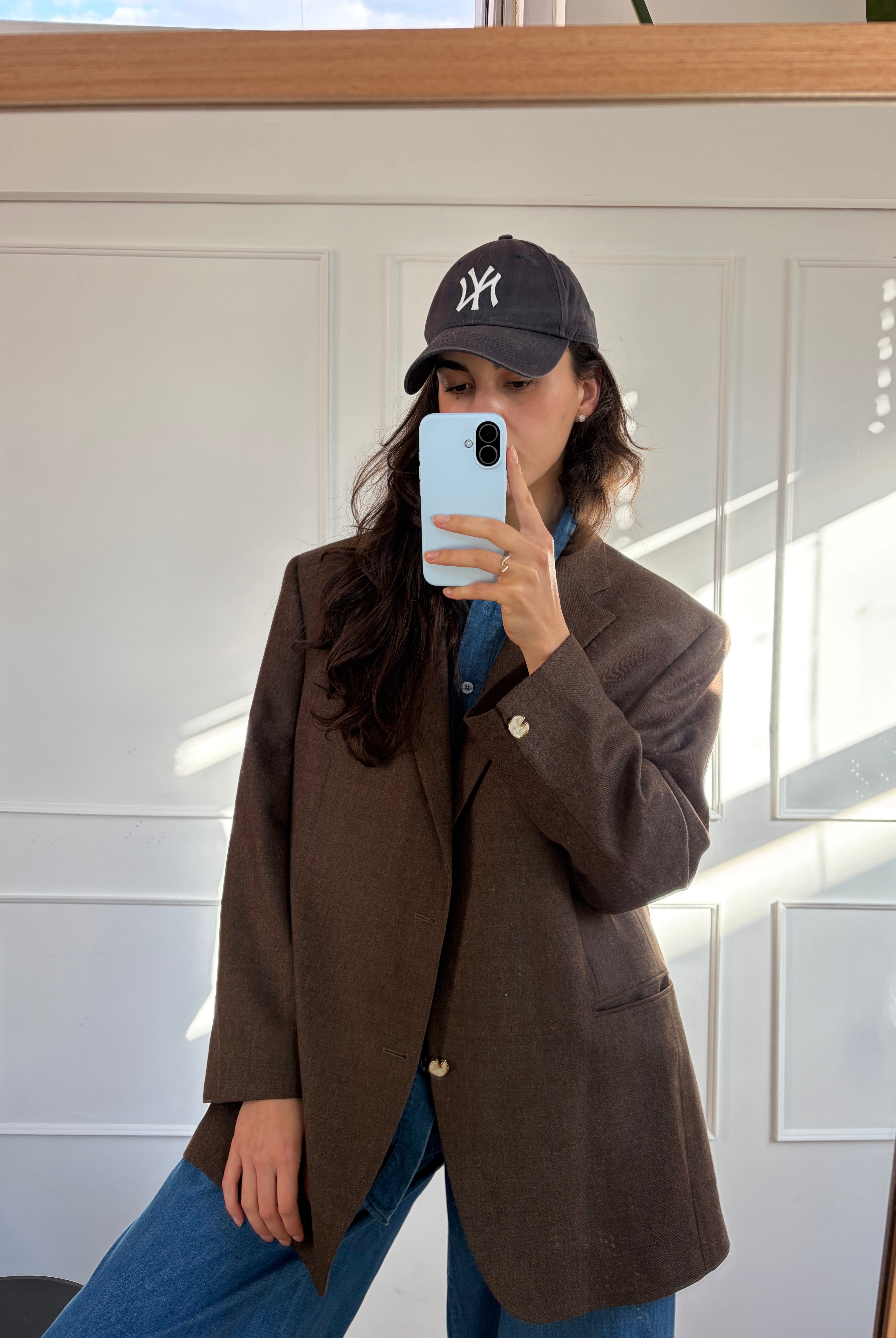 A woman takes a mirror selfie wearing an oversized dark brown blazer over a blue shirt, paired with blue jeans and a black New York Yankees baseball cap.