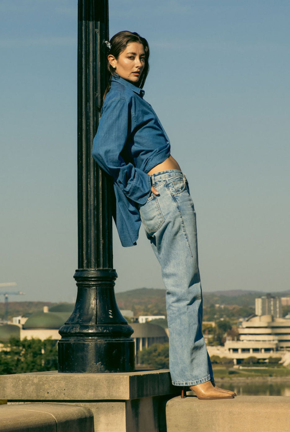 Person in blue shirt and jeans leaning against a lamp post with a cityscape in the background