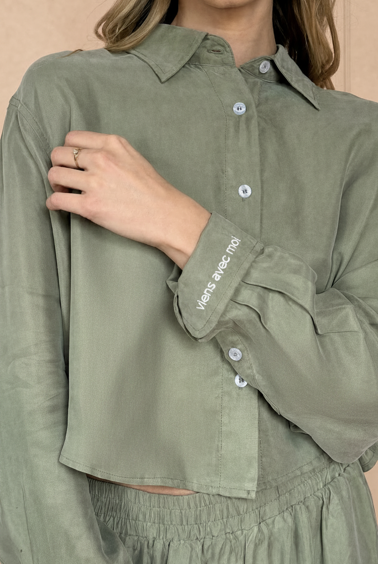 Person wearing a green shirt with rolled-up sleeves, with embroidered viens avec moi on the cuff. The model picture is on a beige background