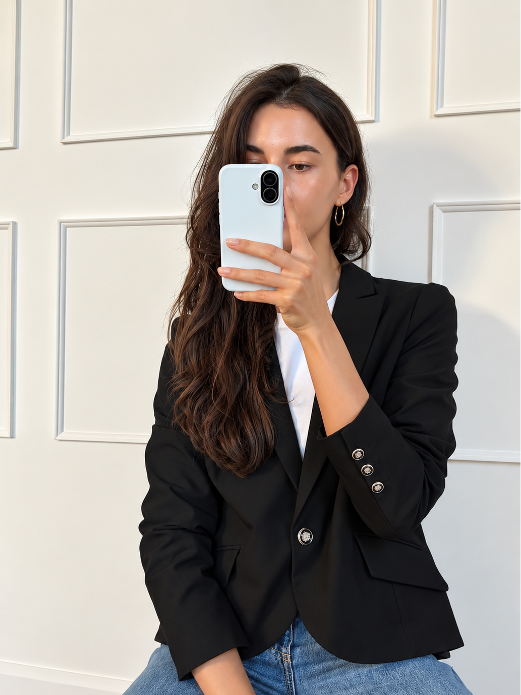 A woman taking a mirror selfie wearing a black blazer over a white t-shirt and blue jeans, holding a white smartphone.