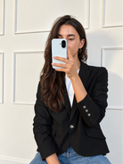 A woman taking a mirror selfie wearing a black blazer over a white t-shirt and blue jeans, holding a white smartphone.
