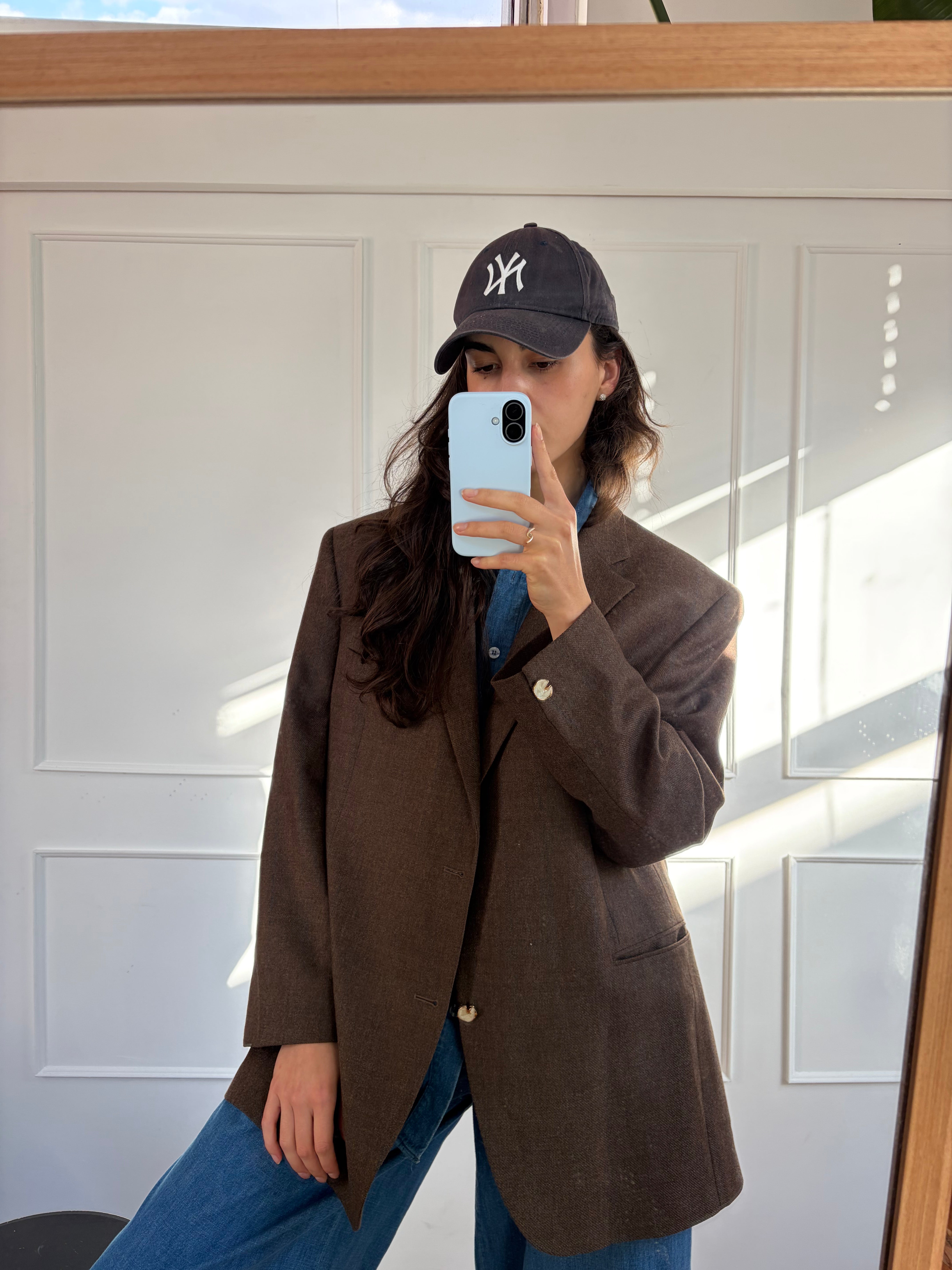 A woman takes a mirror selfie wearing an oversized dark brown blazer over a blue shirt, paired with blue jeans and a black New York Yankees baseball cap.