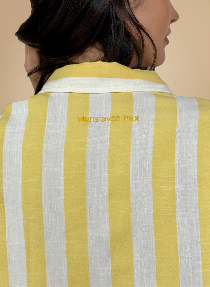 Yellow and white striped shirt with embroidered viens avec moi on the back on a beige background