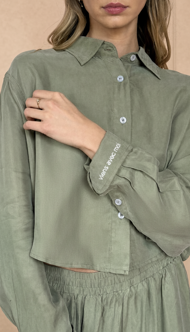 Person wearing a green shirt with rolled-up sleeves, with embroidered viens avec moi on the cuff.  The model picture is on a beige background